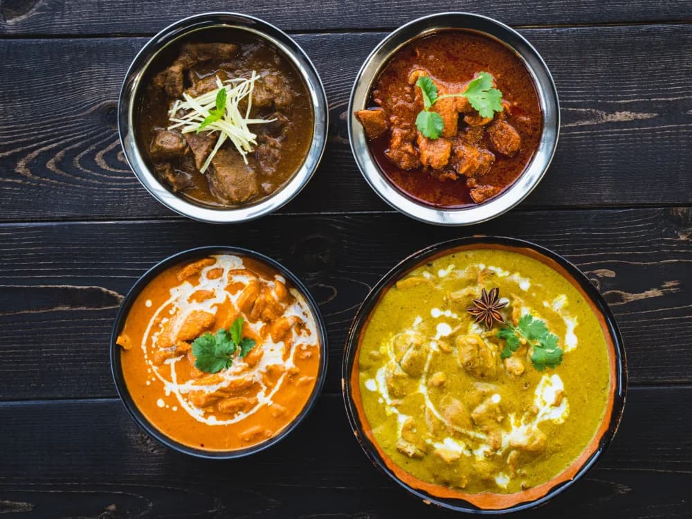 Four Indian dishes in steel bowls on a dark wooden table