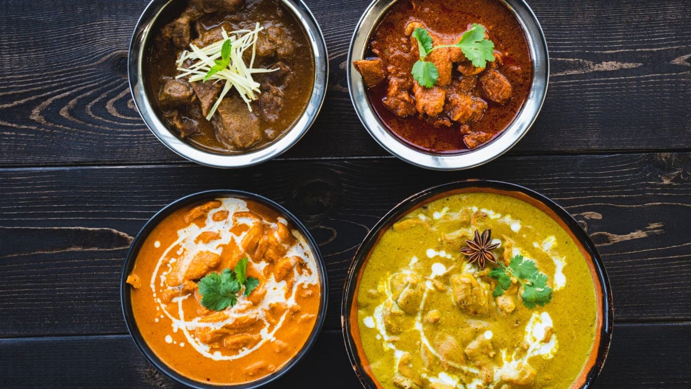 Four Indian dishes in steel bowls on a dark wooden table