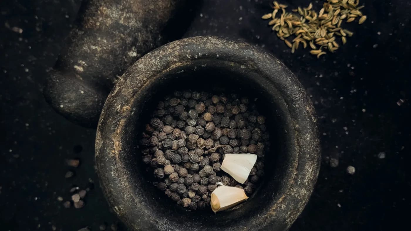 A stone mortar and pestle with whole peppercorns and garlic, seen from above